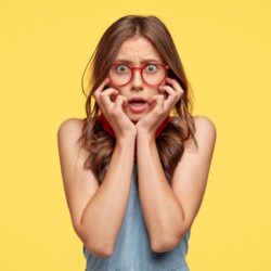concerned-nervous-timid-woman-keeps-hands-cheeks-looks-with-frightened-expression-wears-optical-glasses-feels-worried-as-made-mistake-has-insecure-expression-isolated-yellow-wall_273609-29188